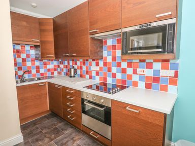 A kitchen with wooden cabinets a tiled backsplash in red blue and light blue colors a built in oven stovetop microwave electric kettle and a double sink at Apartment 2 in Tenby