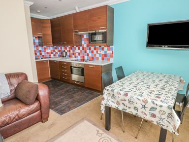 A kitchen and dining area with wooden cabinets a tiled backsplash a dining table with a patterned tablecloth chairs a wall-mounted television and part of a brown leather sofa at Apartment 2 in Tenby