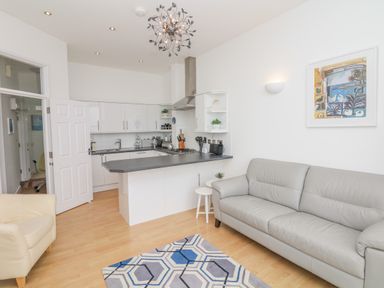A living room with a gray sofa a cream armchair a patterned rug and a kitchen with white cabinets and a black countertop at Oceans Side in Ilfracombe