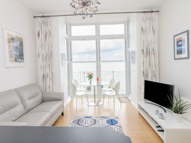 A living room with a gray sofa a round dining table with chairs and a television on a stand at Oceans Side in Ilfracombe