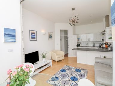 A living room with a sofa armchair rug and TV next to a kitchen with white cabinets and a countertop at Oceans Side in Ilfracombe