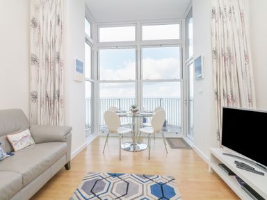 A living room with a grey sofa a glass dining table with four white chairs a television on a white stand and large windows overlooking the sea at Oceans Side Ilfracombe