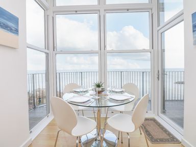 A dining area with a glass table and four white chairs by large windows overlooking the sea at Oceans Side in Ilfracombe