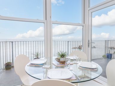 A dining area with a glass round table set for four beside large windows overlooking a balcony and the sea at Oceans Side in Ilfracombe