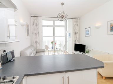 A kitchen with a countertop overlooking a living area with a sofa dining table chairs television and large windows at Oceans Side in Ilfracombe