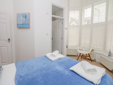 A bedroom with a bed covered by a blue blanket and white towels near a white chair by windows with white shutters at Oceans Side in Ilfracombe