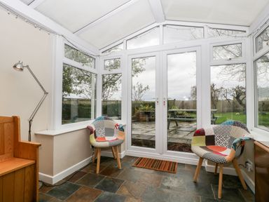 A conservatory with two patterned chairs a wooden bench a floor lamp and glass doors leading to a garden at Pippins in Bream