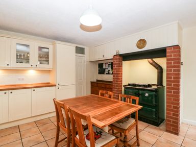 A kitchen with wooden table and chairs a green stove in a brick alcove and white cabinets at Pippins in Bream