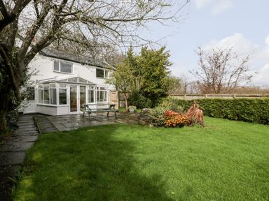A backyard with a grassy lawn a paved patio area a wooden picnic table bushes and trees near a white house with a conservatory at Pippins in Bream