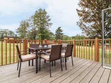 An outdoor wooden deck with a dining table and six chairs with cushions overlooking a lawn and trees at Ash in Truro