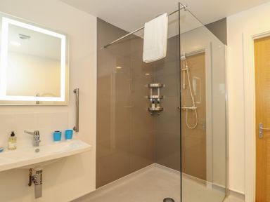 A bathroom with a wall-mounted sink a mirror two toothbrush holders a shower area with shelves and a towel hanging at Ash in Truro