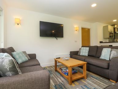 A living room with two gray sofas a wooden coffee table a wall-mounted television and a kitchen area at Ash in Truro