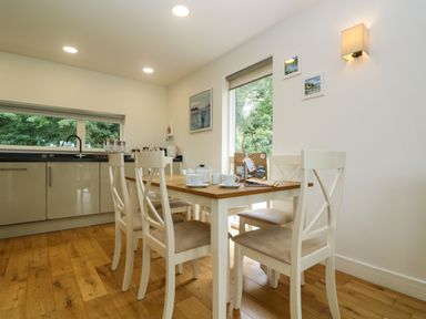 A dining area with a wooden table and four white chairs next to a kitchen with cabinets and a window at Ash in Truro