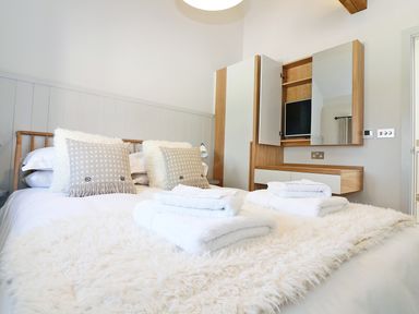 A bedroom with a bed with pillows and folded towels with a wall-mounted cabinet with a TV at Long House Boskensoe Barns in Mawnan Smith
