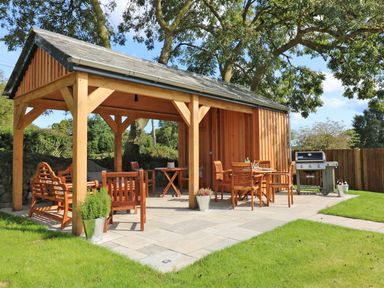 A wooden outdoor gazebo with chairs tables grill and grass at Long House Boskensoe Barns in Mawnan Smith