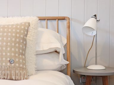 A wooden bed frame with white pillows and a beige polka dot cushion next to a table with a white lamp at Long House Boskensoe Barns in Mawnan Smith