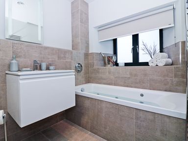 A bathroom with a bathtub a white vanity with soap dispensers and a window with rolled shades at Long House Boskensoe Barns in Mawnan Smith