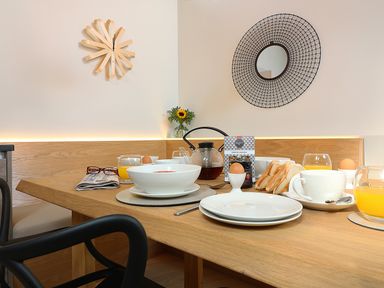 A dining table set with bowls, plates, soft boiled eggs, toast, glasses of orange juice, a teapot, and a box of granola in a dining area with wall clock and mirror at Long House Boskensoe Barns in Mawnan Smith