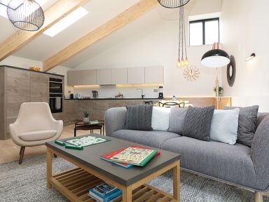 A living area with a gray sofa with cushions a beige armchair a wooden coffee table with books and a board game and a kitchen in the background at Long House Boskensoe Barns in Mawnan Smith