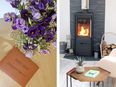 A bouquet of purple flowers beside a guest book on a wooden table and a room with a wood-burning stove a basket of firewood and a small table with a plant and a book at Long House Boskensoe Barns in Mawnan Smith