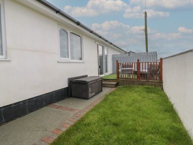 A side yard with grass a paved path a storage box and a raised deck with seating at Sea Spray in Dinas Dinlle