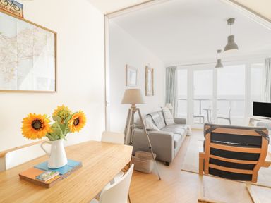 A living room with a wooden dining table and sunflowers in a vase next to a gray sofa and armchair with large windows overlooking the sea at 12 West End Point in Pwllheli