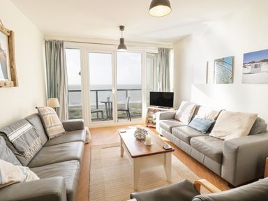 A living room with two sofas, a coffee table, a television on a stand, and a balcony with a table and two chairs overlooking the ocean at 12 West End Point in Pwllheli