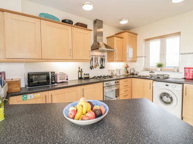 A kitchen with wooden cabinets a washing machine a bowl of fruit on the counter and kitchen appliances at 12 West End Point in Pwllheli