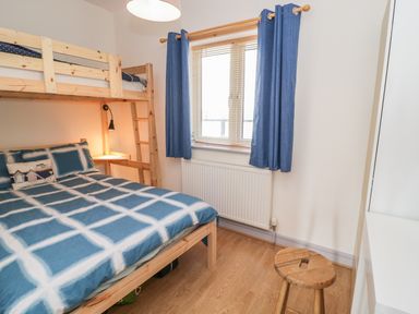A bedroom with a wooden bunk bed with blue and white checkered bedding a window with blue curtains a wooden stool and wood flooring at 12 West End Point Pwllheli