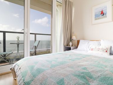 A bedroom with a double bed next to a balcony with chairs and a table overlooking the sea at 12 West End Point in Pwllheli