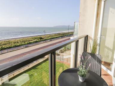 A balcony with a round black table and a chair overlooking a beach and a road at 12 West End Point in Pwllheli