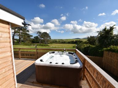 An outdoor wooden deck with a hot tub filled with bubbles and a bottle with glasses overlooking fields and trees at Y Caban Clyd in Morfa Nefyn