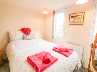 A bedroom with a double bed with white bedding and red towels with a heart shaped pillow a wooden nightstand a framed picture on the wall and two windows at Y Caban Clyd in Morfa Nefyn