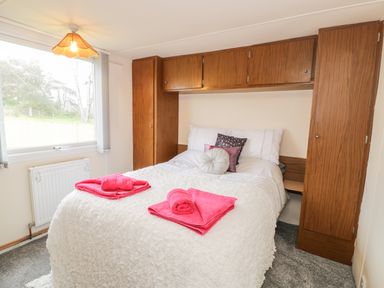 A bedroom with a double bed with white bedding and pink towels on it wooden cabinets around the bed and a window with a view of trees at Y Caban Clyd in Morfa Nefyn
