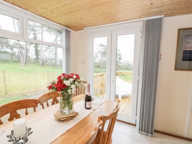 A dining room with a wooden table and chairs a vase of flowers a bottle of champagne and two glasses at Y Caban Clyd in Morfa Nefyn