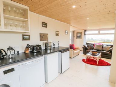 A kitchen area with black countertop appliances and a living room with sofas and a red rug at Y Caban Clyd in Morfa Nefyn
