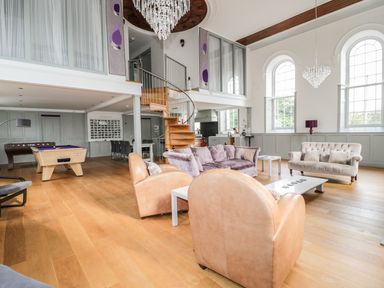 A spacious living room with beige leather chairs purple sofa two large arched windows and a wooden spiral staircase at Capel Bryn Bachau in Chwilog near Criccieth