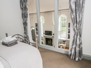 A bedroom with a metal bed frame folded towels and large windows looking into a living room with a fireplace and television at Capel Bryn Bachau in Chwilog near Criccieth