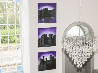 Three paintings of castles with purple skies on a white wall near a window and a hanging chandelier at Capel Bryn Bachau in Chwilog near Criccieth