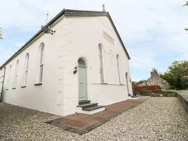 Exterior view of a white building with arched windows and doors surrounded by gravel and a brick pathway with another stone building in the background at Capel Bryn Bachau Chwilog near Criccieth