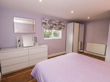 A bedroom with a bed a white dresser with mirrors a window with floral blinds a wardrobe and a radiator at Nant Y Felin Lodge in Aberdaron
