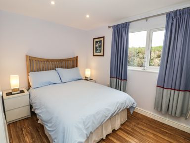 A bedroom with a wooden bed frame covered in light blue bedding two white nightstands with lamps and blue curtains over a window at Nant Y Felin Lodge in Aberdaron