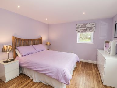 A bedroom with a wooden bed, lavender bedding, two bedside tables with lamps, a window with floral blinds, and a white dresser at Nant Y Felin Lodge in Aberdaron