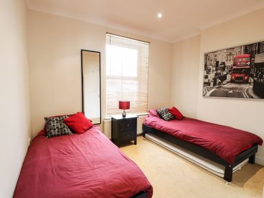 A bedroom with two single beds with red bedding a black nightstand with a lamp a tall mirror and a black and white cityscape picture with a red bus at Apartment 3 Marian Y Mor in Pwllheli