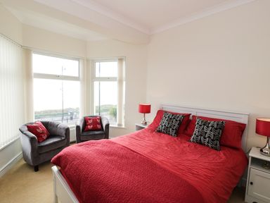 A bedroom with a double bed covered in red bedding flanked by two bedside tables with red lamps and two gray chairs with red cushions by the windows at Apartment 3 Marian Y Mor in Pwllheli
