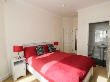 A bedroom with a double bed with red bedding two bedside tables with lamps and an ensuite bathroom at Apartment 3 Marian Y Mor in Pwllheli