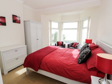 A bedroom with a red bedspread two black and white pillows two black chairs with red pillows a white wardrobe and chest of drawers at Apartment 3 Marian Y Mor in Pwllheli