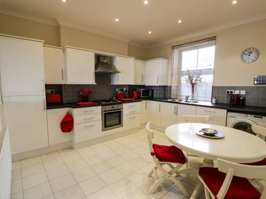 A kitchen with white cabinets black countertops a round dining table with red cushioned chairs and appliances at Apartment 3 Marian Y Mor Pwllheli