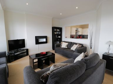 A living room with grey sofas a black coffee table a tv and a doorway to a kitchen at Apartment 3 Marian Y Mor in Pwllheli