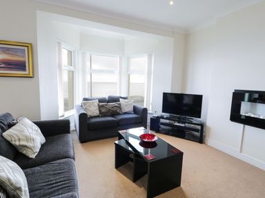 A living room with two gray sofas a black coffee table with a red bowl a television and a view of the sea through large windows at Apartment 3 Marian Y Mor in Pwllheli
